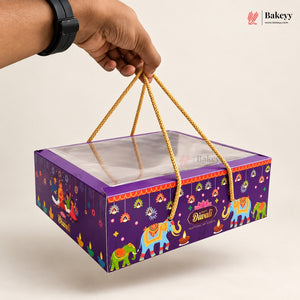 A hand holds a purple Diwali-themed gift box with a clear window and gold rope handles. The box is decorated with colorful elephants, hanging lanterns, and festive patterns, with the brand name Bakeyy visible in the top right corner.