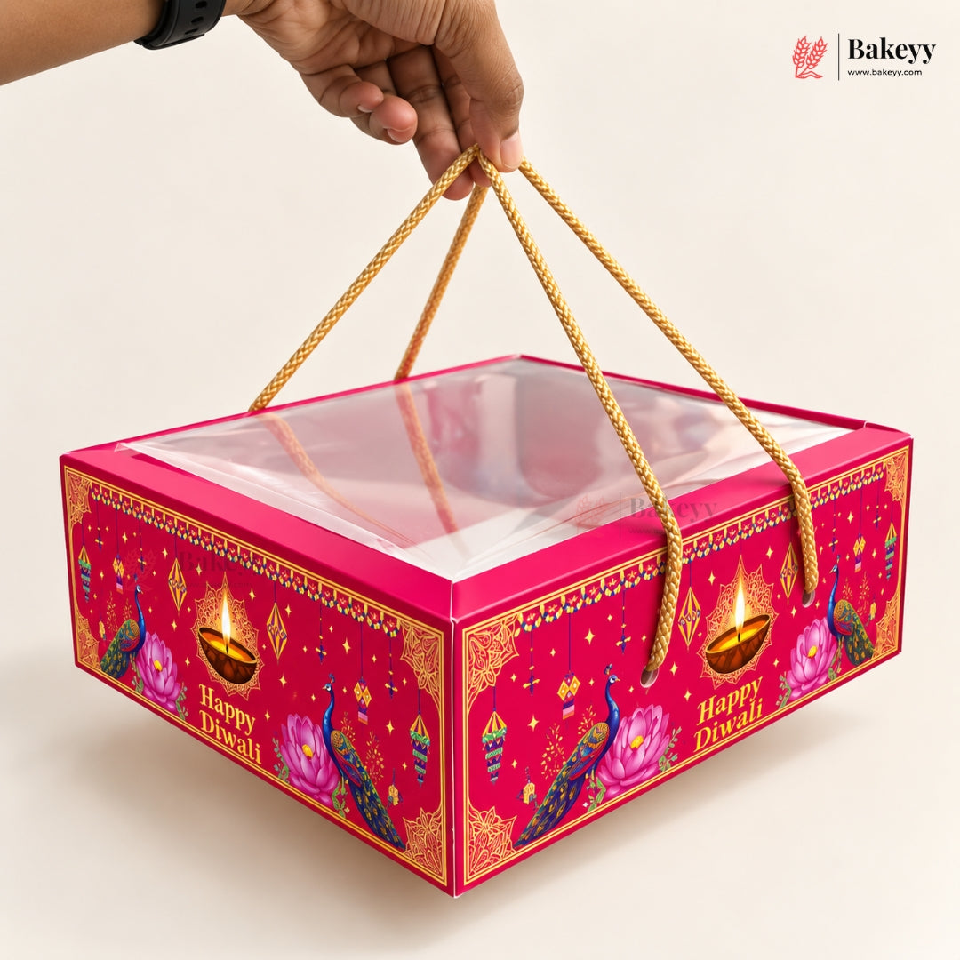A hand holds a bright pink Diwali-themed gift box with a clear plastic window on top and gold rope handles. The box is decorated with peacocks, lotus flowers, and oil lamps, with 'Happy Diwali' printed on the front.