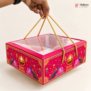 A hand holds a bright pink Diwali-themed gift box with a clear plastic window on top and gold rope handles. The box is decorated with peacocks, lotus flowers, and oil lamps, with 'Happy Diwali' printed on the front.