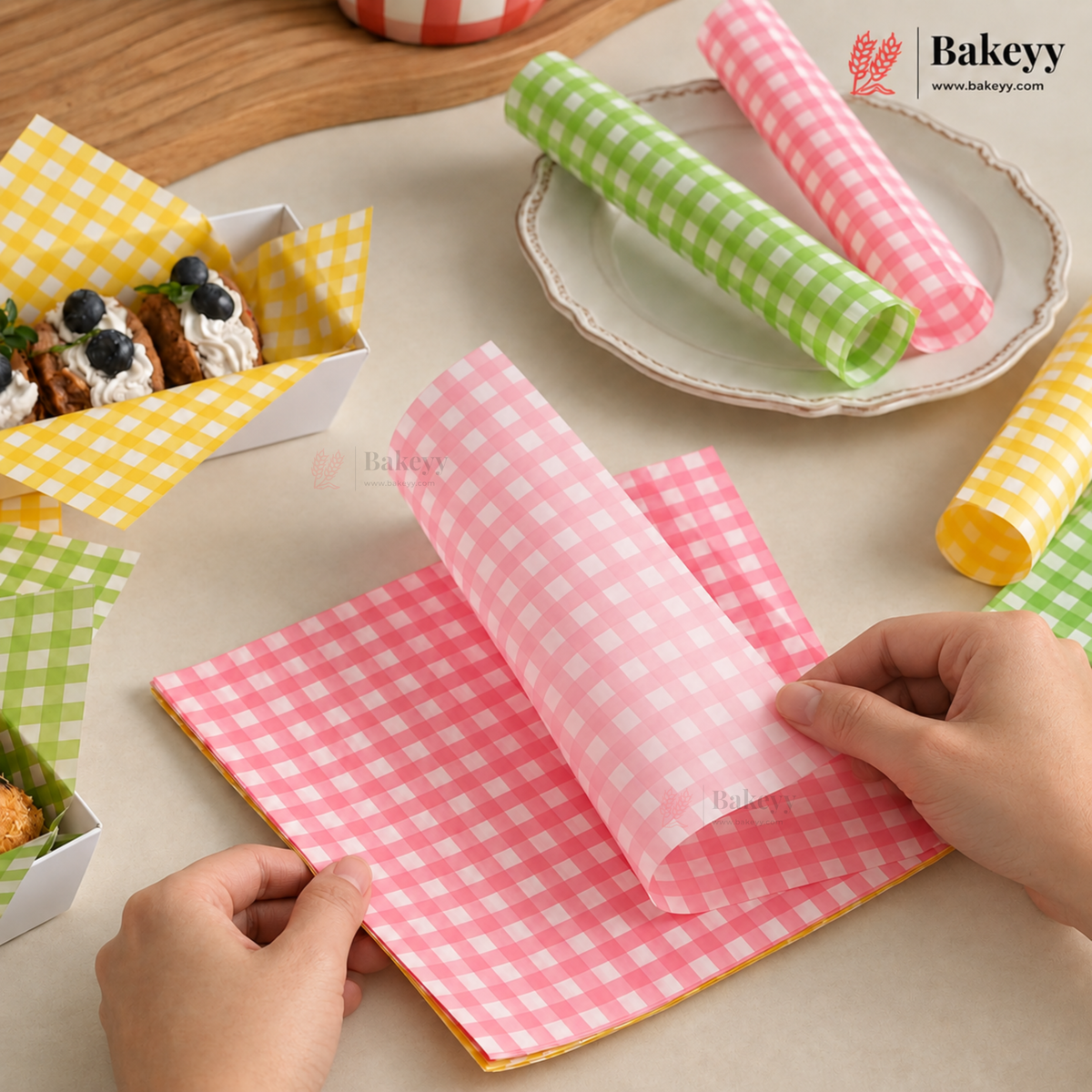 Pink and white checkered baking parchment paper being held open by hands, with other checkered parchment rolls and trays in the background on a beige tabletop.