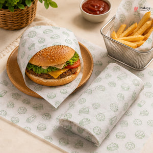 A gourmet hamburger wrapped in branded white paper sleeve with green burger illustrations, served on a wooden plate next to french fries and ketchup, with a matching folded sleeve in the foreground.