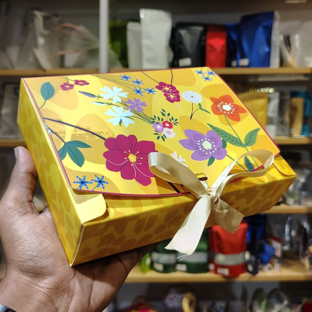 A hand holds a yellow gift box decorated with a colorful floral pattern and tied with a cream ribbon, set against a blurred background of shelves with bags.