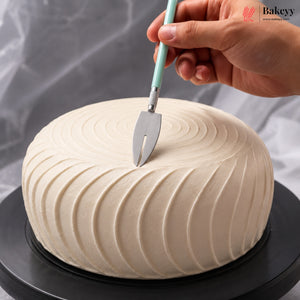 A hand holds a cake server above a round, cream-colored frosted cake on a black rotating stand, with a gray draped fabric background. A 'Bakeyy' watermark is visible in the top right corner.