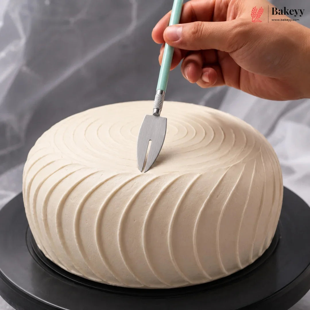 A hand holds a cake server above a round, cream-colored frosted cake on a black rotating stand, with a gray draped fabric background. A 'Bakeyy' watermark is visible in the top right corner.
