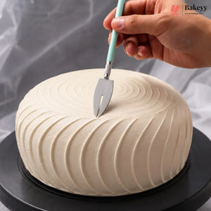A hand holds a cake server above a round, cream-colored frosted cake on a black rotating stand, with a gray draped fabric background. A 'Bakeyy' watermark is visible in the top right corner.