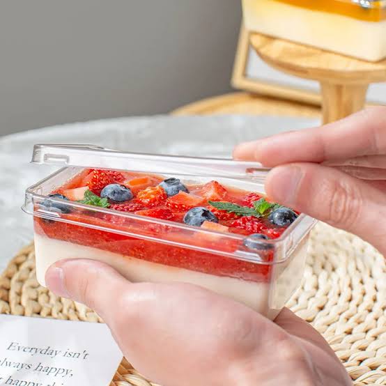 Hand holding a container of dessert with strawberries and blueberries on a textured surface.