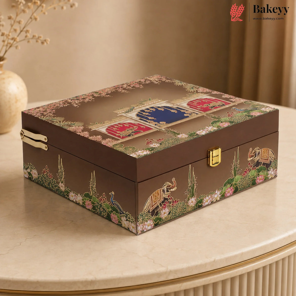 A rectangular decorative jewelry box with a dark brown base and intricate multicolor printed illustrations of peacocks, elephants, flowers, and traditional motifs on the lid and sides. The box has gold-tone metal latches and is displayed on a beige marble table with a warm, elegant background.