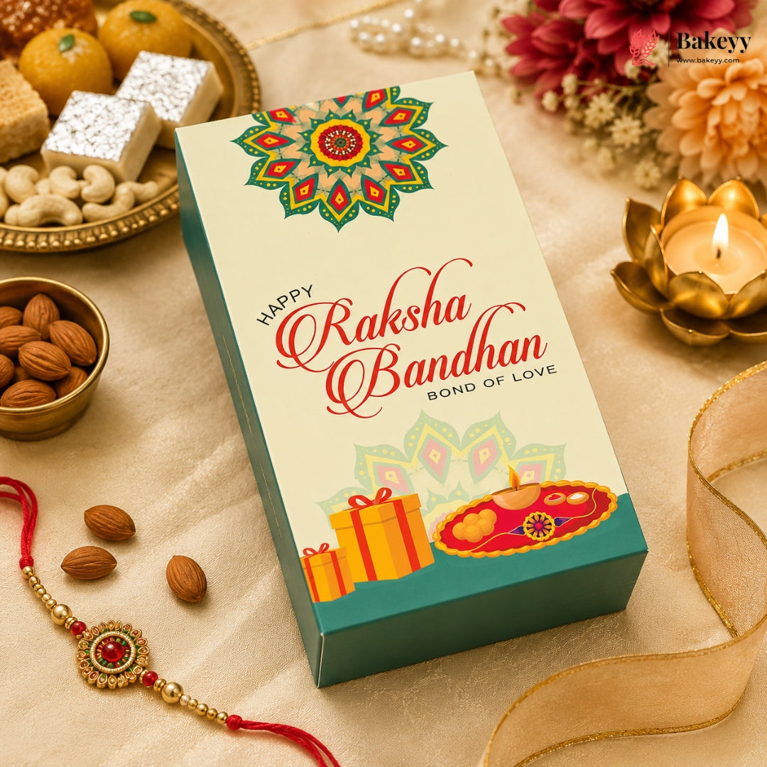 A rectangular gift box for Raksha Bandhan sits on a beige fabric surface surrounded by festive props, including almonds, a red and gold Rakhi, sweets, flowers, a lit lotus candle, and a gold ribbon. The box front is cream with a colorful mandala design, red script reading 'Happy Raksha Bandhan,' and illustrations of gift boxes and a diya.