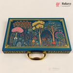A rectangular blue decorative gift box with a gold handle, featuring an illustrated forest scene with colorful trees and floral motifs on the lid, photographed on a light beige background. The Bakeyy logo appears in the top right corner.