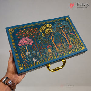 A hand holds a rectangular decorative gift box with a blue background and an intricate multicolor forest illustration featuring trees, flowers, and mushrooms, accented with gold trim and a gold handle. The Bakeey brand logo is visible in the top right corner.