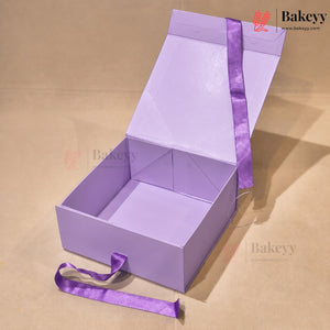 A lavender purple cardboard gift box with its lid propped open, featuring a matching purple satin ribbon tied around it and a loose end resting on the surface. The box is photographed on a neutral beige background with a Bakeyy watermark.