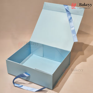 A light blue cardboard gift box with an open hinged lid, a matching blue ribbon tied around the package, and a blue ribbon loop handle, shown on a beige background.