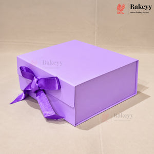 A lavender purple rectangular gift box with a matching purple satin ribbon bow, set against a neutral beige background. The Bakeyy brand logo is visible in the top right corner.