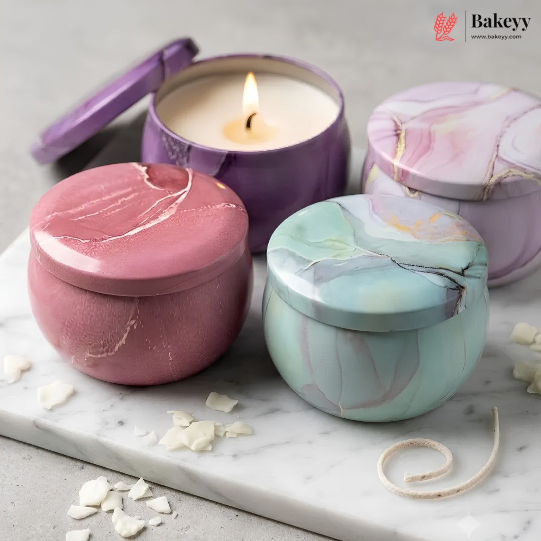 Three marbled metal candle tins with matching lids arranged on a white marble board. One tin is open and lit, showing cream wax and a small flame.