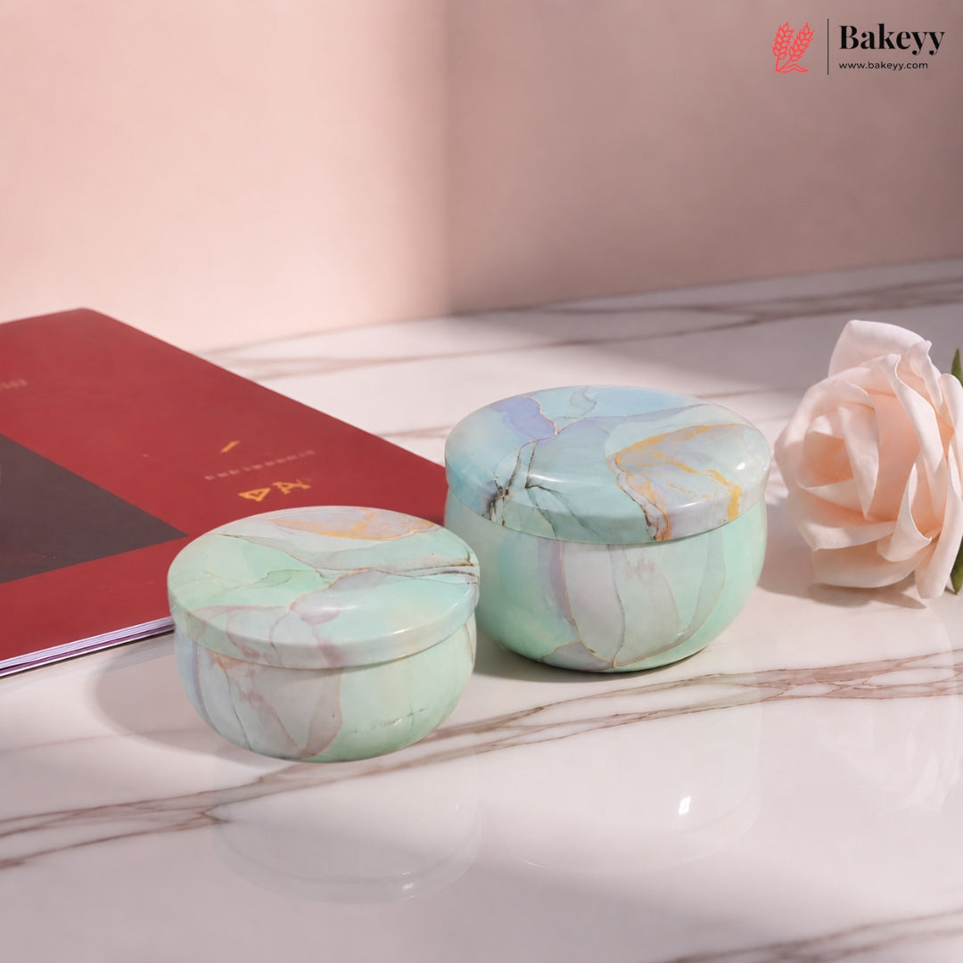 Two round lidded jars with a light green marble pattern are displayed on a white marble surface next to a red book and a pale pink rose. The background is a soft beige wall.