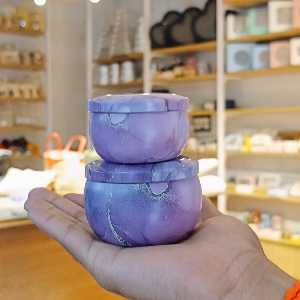 A hand holding two stacked round purple marbled containers with matching lids in a softly blurred retail shop setting.