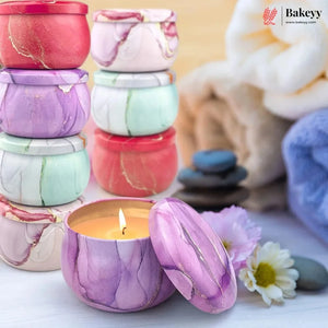 A set of marble-patterned candle tins with matching lids, shown with one tin open and lit in the foreground. The tins come in multiple colors including purple, pink, red, mint green, and white, with a spa-themed background featuring folded towels, stones, and flowers.