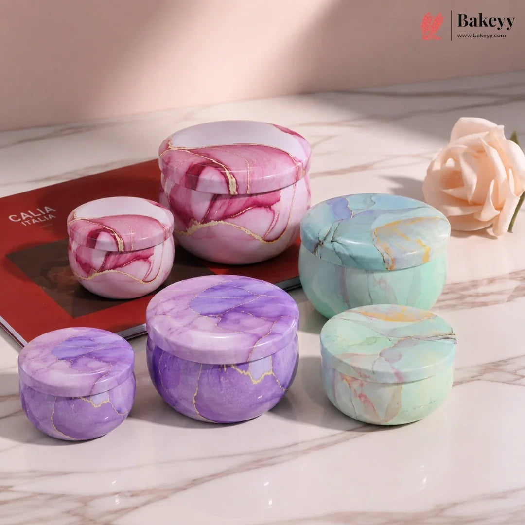 Six round marble-patterned cosmetic jars with matching lids are arranged on a light marble surface. The jars come in pink, purple, and mint green marble designs with thin gold veining.