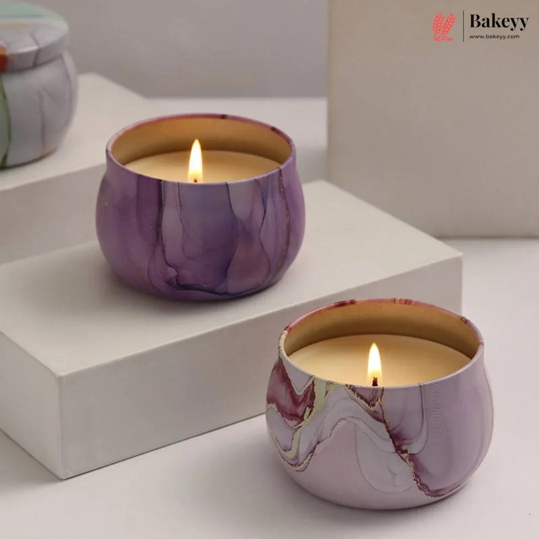 Two lit marbled candle holders in purple, lavender, and gold tones, styled on white display blocks with a neutral background. A Bakeyy logo watermark is visible in the top right corner.