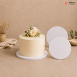 A cream-colored frosted cake with white flowers and green leaves sits on a round white cake stand, with two blank white round cake stands displayed beside it on a beige background. The Bakeyy logo is visible in the top right corner.