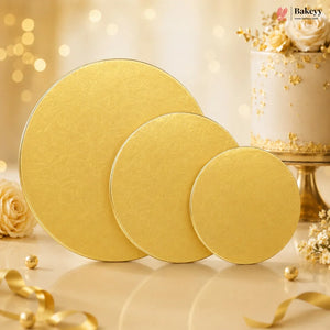 Three round gold cake stands of decreasing sizes arranged on a light surface, with a decorated white cake and gold floral accents in the background. The Bakeyy logo appears in the top right corner.