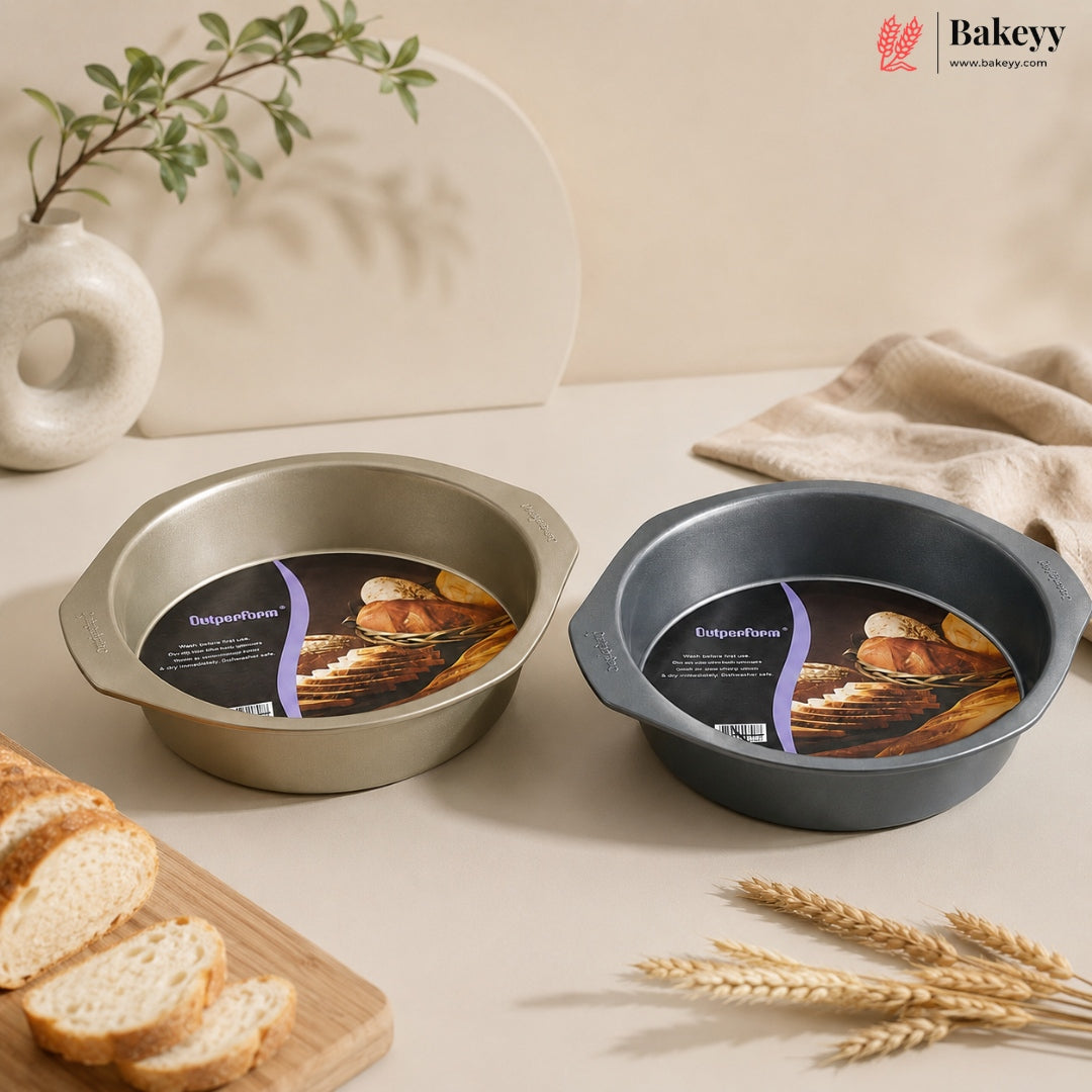 Two round baking pans with handles, one brushed silver and one dark gray, both still attached to their product labels, displayed on a beige tabletop with bread, wheat, and a ceramic vase in the background.