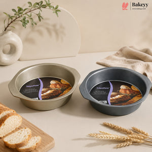 Two round baking pans with handles, one brushed silver and one dark gray, both still attached to their product labels, displayed on a beige tabletop with bread, wheat, and a ceramic vase in the background.