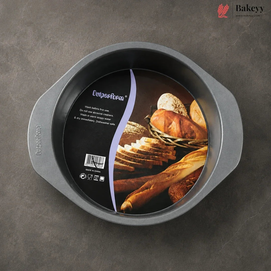 A round non-stick baking pan with side handles, shown from above on a dark gray textured surface. The inside of the pan has a large black product label featuring bread imagery and the brand name 'Cupcake Pan'.