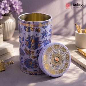 A decorative cylindrical metal tea tin with a matching lid, featuring a lavender and gold floral pattern with Chinese characters, displayed on a styled desk surface.