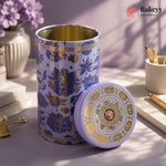 A decorative cylindrical metal tea tin with a matching lid, featuring a lavender and gold floral pattern with Chinese characters, displayed on a styled desk surface.
