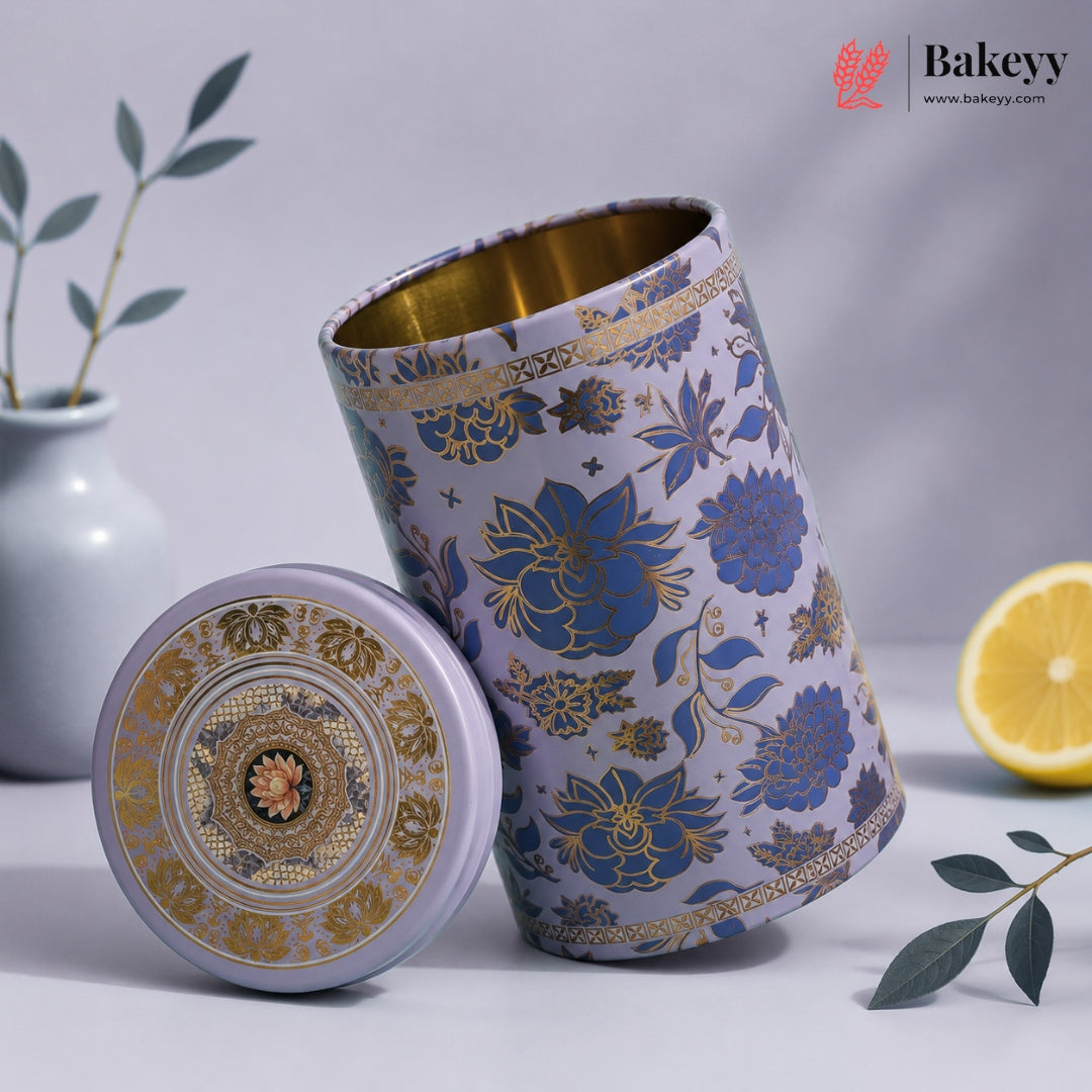 A decorative cylindrical metal tea or coffee canister with its lid placed beside it. The canister has a lavender-purple exterior with blue floral motifs and gold detailing, and a gold interior.