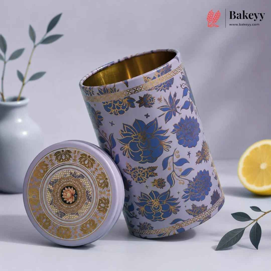 A decorative cylindrical metal tea or coffee canister with its lid placed beside it. The canister has a lavender-purple exterior with blue floral motifs and gold detailing, and a gold interior.