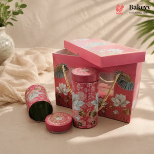 A pink floral bakery gift set featuring a rectangular box with a hinged lid, a cylindrical tin with rope handles, a small round tin, and an open tin showing green contents, styled on a beige fabric surface with soft shadows and a Bakeyy logo in the top right corner.