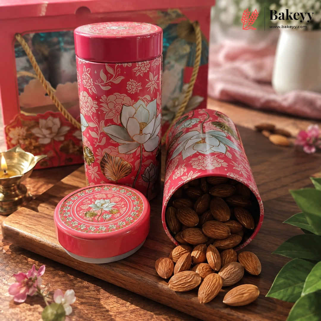 A pink floral metal tin container for baking or tea, shown closed and open with almonds spilling out on a wooden surface. A matching gift box is visible in the background, along with decorative flowers, a small brass lamp, and green leaves.