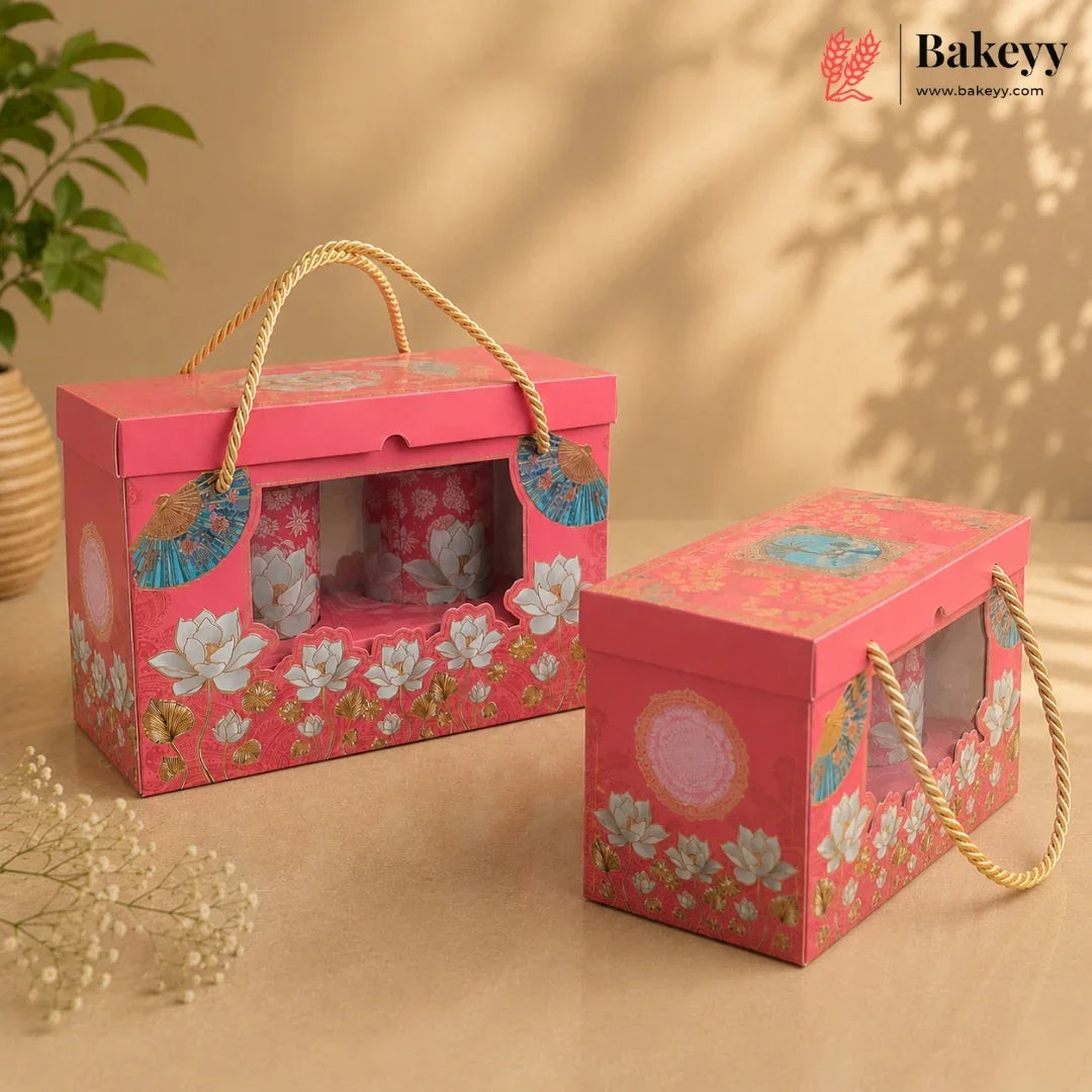 Two pink decorative bakery gift boxes with floral prints and rope handles, shown on a warm beige background with a plant and dried flowers as props. The Bakeyy logo appears in the top right corner.