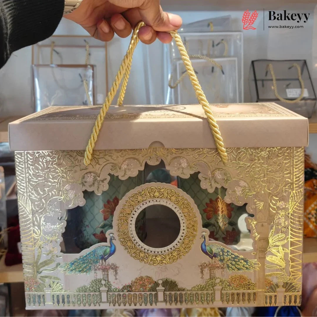 An ornate rectangular gift box with a lid, decorated with gold foil patterns, peacock illustrations, and a central oval window. The box has twisted rope handles and is being held by a hand in a retail setting.