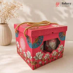 A pink decorative gift box with a rope handle, floral and fan pattern, and a clear window showing a pink lotus cup inside. A beige vase with dried stems sits in the background on a warm cream surface.