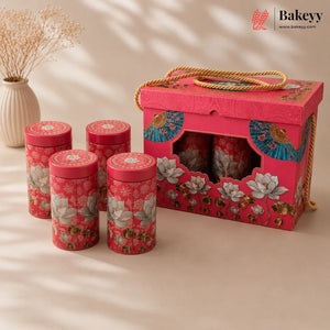 A pink tea gift set with four cylindrical floral tins and a matching decorative box, styled on a beige surface with a vase of dried flowers in the background. The Bakeyy logo is visible in the top right corner.
