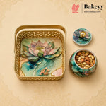 A decorative gold metal gift tray with a lotus pond design, shown alongside a matching lidded container and a small jar of nuts. The Bakeyy brand logo is visible in the top right corner.