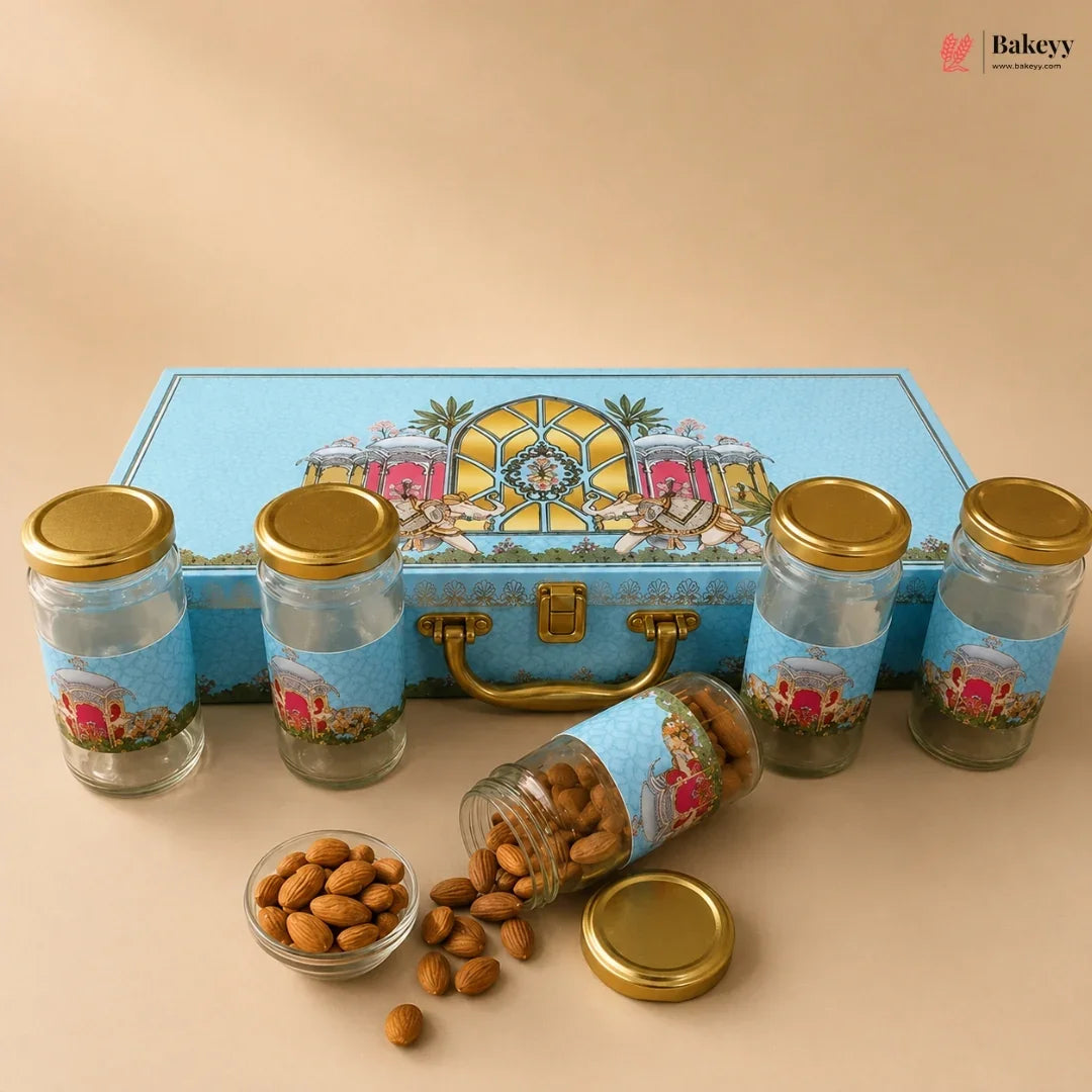 A gourmet gift set of five glass jars with gold lids and blue decorative labels, arranged in front of a matching blue illustrated storage box. One jar is tipped over with almonds spilling out onto a beige surface, with a small glass dish of almonds in the foreground.