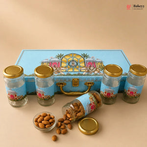 A gourmet gift set of five glass jars with gold lids and blue decorative labels, arranged in front of a matching blue illustrated storage box. One jar is tipped over with almonds spilling out onto a beige surface, with a small glass dish of almonds in the foreground.