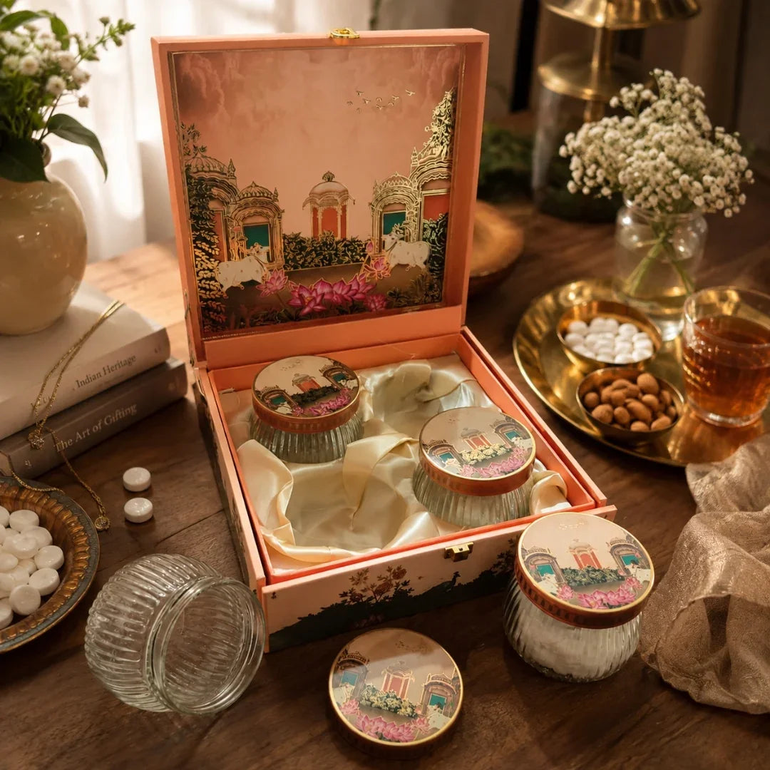 A decorative open gift box containing several small round jars with illustrated lids, arranged on a wooden table with flowers, books, and a glass of tea nearby.