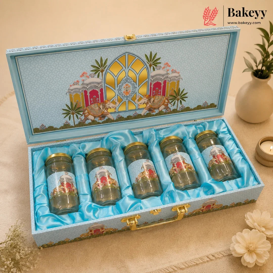 An open decorative gift box containing five small glass jars with gold lids, set on a beige surface with flowers and a plant in the background. The box lid is decorated with a colorful architectural illustration, and the interior is lined with light blue satin.