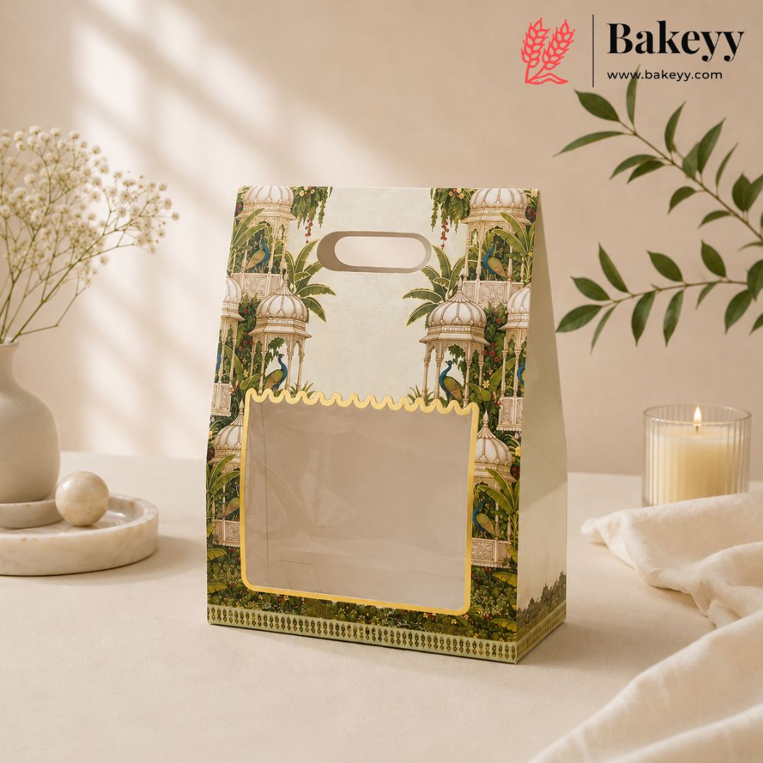 A decorative paper gift bag with a tropical pavilion print and a clear window on the front, styled on a tabletop with dried flowers, a candle, and a leafy branch in a warm neutral setting. The Bakeyy logo appears in the top right corner.