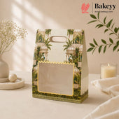 A decorative paper gift bag with a tropical pavilion print and a clear window on the front, styled on a tabletop with dried flowers, a candle, and a leafy branch in a warm neutral setting. The Bakeyy logo appears in the top right corner.
