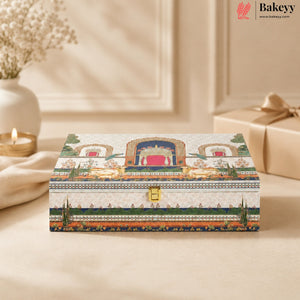 A rectangular decorative gift box with an ornate traditional-style illustration on the lid, featuring a gold clasp and a small Bakeyy logo in the top right corner. The box is styled on a beige surface with a blurred background showing a candle, a vase, and another wrapped gift.