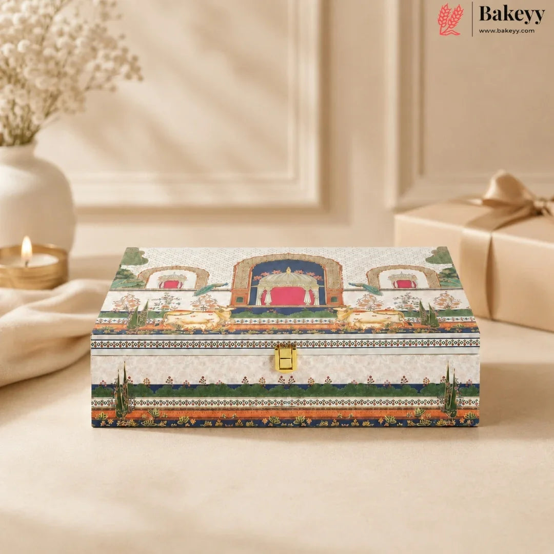 A rectangular decorative gift box with an ornate traditional-style illustration on the lid, featuring a gold clasp and a small Bakeyy logo in the top right corner. The box is styled on a beige surface with a blurred background showing a candle, a vase, and another wrapped gift.