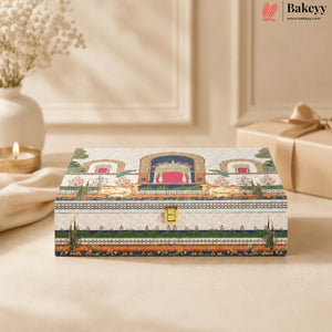 A rectangular decorative gift box with an ornate traditional-style illustration on the lid, featuring a gold clasp and a small Bakeyy logo in the top right corner. The box is styled on a beige surface with a blurred background showing a candle, a vase, and another wrapped gift.