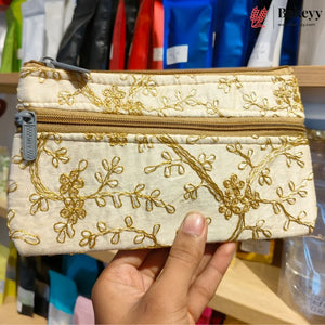 A hand holding a small zippered pouch with gold floral embroidery on a cream fabric background, set against a blurred store shelf backdrop.