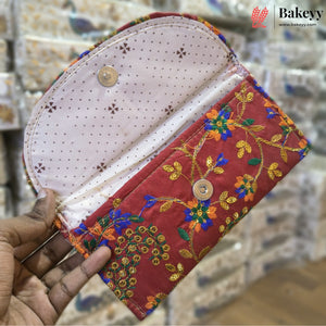 A handmade fabric clutch wallet held open by a hand, featuring a red floral embroidered exterior, a light pink dotted interior lining, and gold-tone snap buttons. The background shows a blurred tiled surface with a Bakeyy watermark logo in the top right.